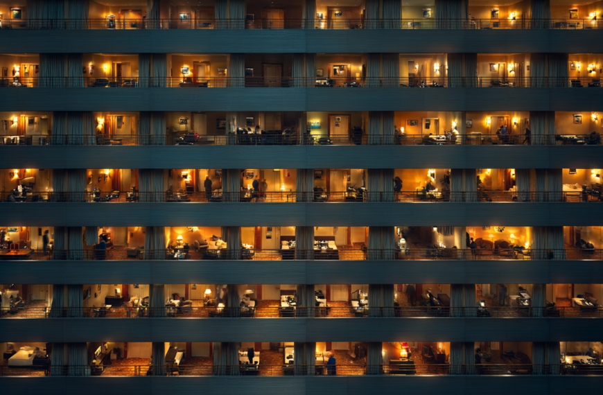 The image shows the exterior of a multi-story building at night, with several floors visible. Each floor has multiple rooms with large windows, through which warm, yellow lighting can be seen. The rooms appear to be hotel rooms or apartments, with visible furniture such as beds, tables, and chairs. Some rooms have people inside, engaged in various activities. The overall atmosphere is cozy and illuminated.