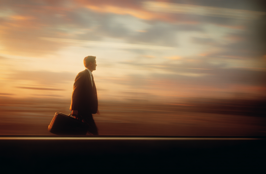 A man in a suit is walking briskly while carrying a briefcase. The background is a blurred sunset sky, creating a sense of motion and urgency.