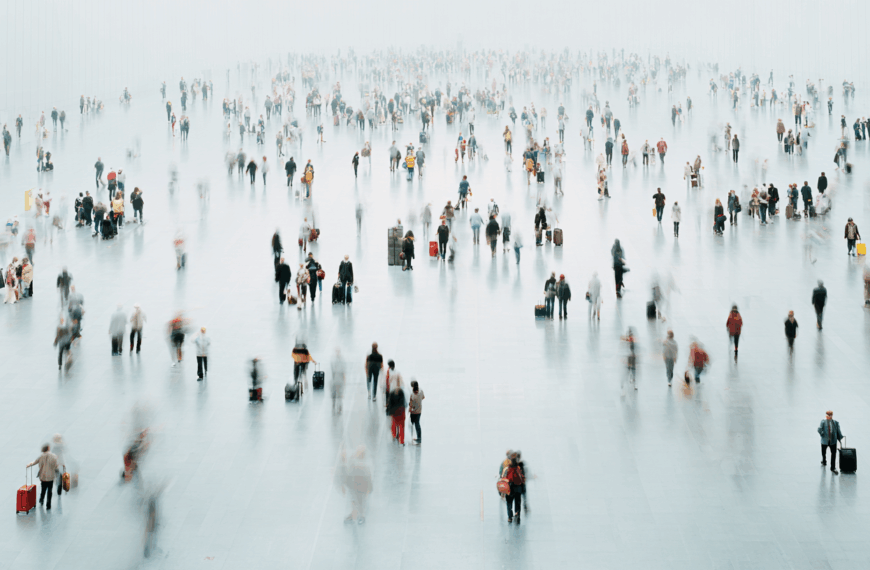 Blurred image of a large, open indoor space with many people walking in various directions. Some individuals are carrying luggage, suggesting a busy transportation hub like an airport or train station. The floor is reflective, and the background is bright and slightly foggy, adding to the sense of motion and activity.