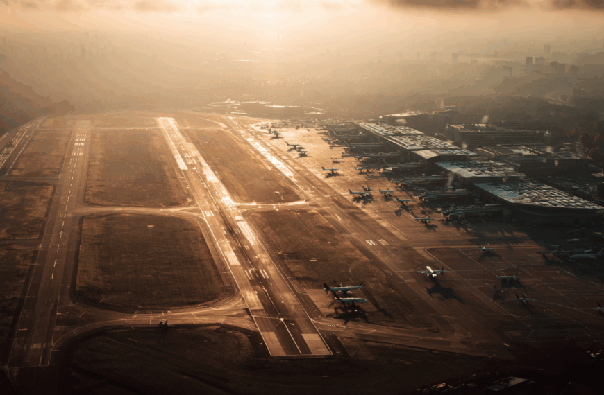 Aerial view of an airport at sunrise or sunset, with multiple runways and taxiways visible. Several airplanes are parked near the terminal buildings on the right side. The scene is bathed in a warm, golden light, with the city skyline faintly visible in the background through a layer of mist or haze.