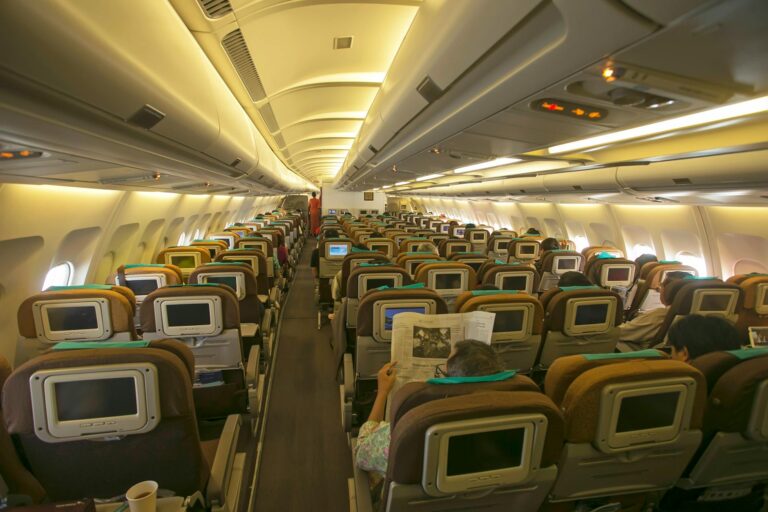 The image shows the interior of an airplane cabin, specifically the economy class section. The seats are arranged in rows, each equipped with personal entertainment screens on the back. Passengers are seated, with some reading newspapers. The cabin is well-lit, and overhead compartments are visible above the seats.