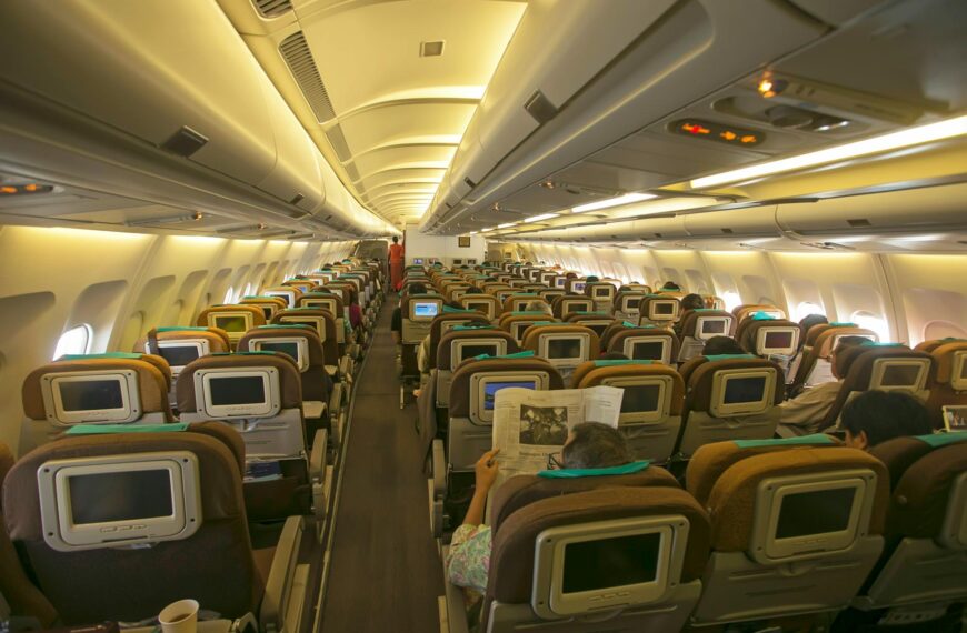 The image shows the interior of an airplane cabin, specifically the economy class section. The seats are arranged in rows, each equipped with personal entertainment screens on the back. Passengers are seated, with some reading newspapers. The cabin is well-lit, and overhead compartments are visible above the seats.