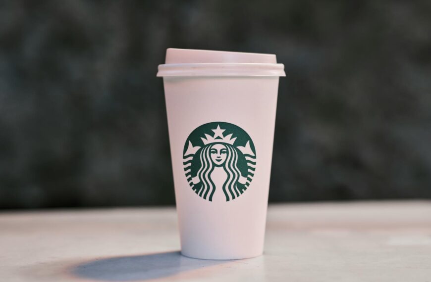 A white disposable coffee cup with a lid, featuring the Starbucks logo, is placed on a surface with a blurred dark background.