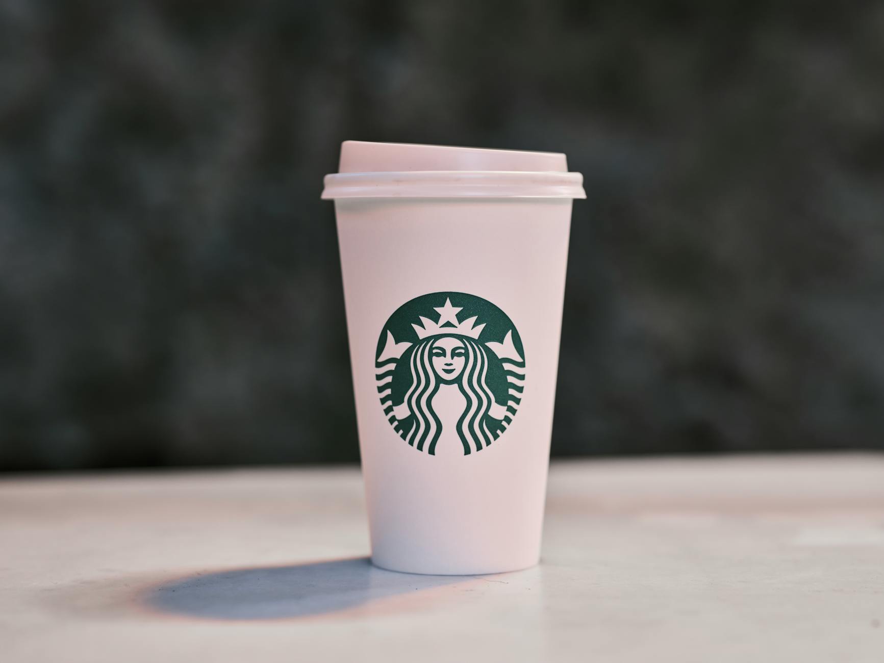 A white disposable coffee cup with a lid, featuring the Starbucks logo, is placed on a surface with a blurred dark background.