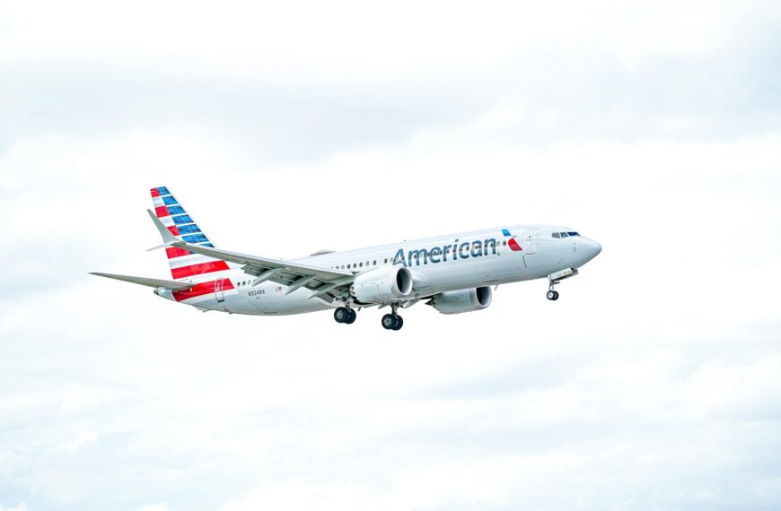 A commercial airplane in flight against a cloudy sky. The aircraft has "American" written on the side and features a red, white, and blue striped tail. The landing gear is visible, indicating it is either taking off or preparing to land.