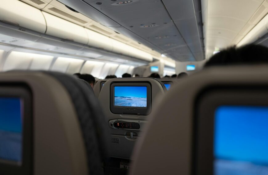 The image shows the interior of an airplane cabin, focusing on the back of several passenger seats. Each seat has a personal entertainment screen displaying an image of a snowy landscape. The cabin is well-lit, and the ceiling and overhead compartments are visible.