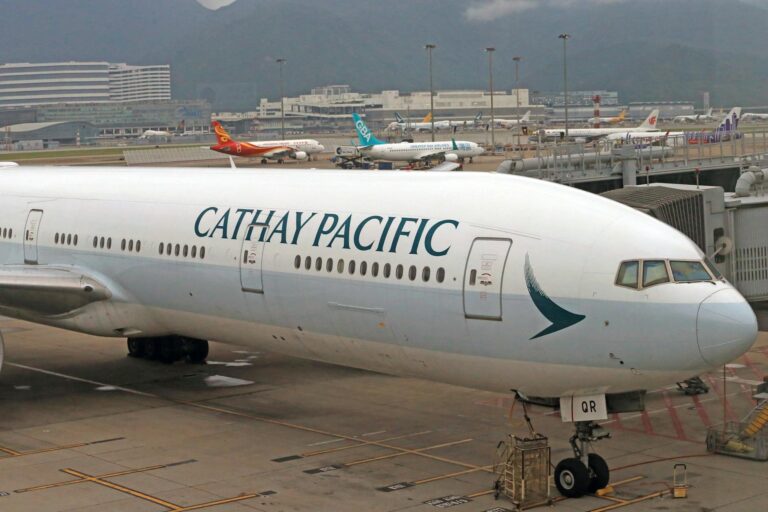 A Cathay Pacific airplane is parked at an airport gate. The aircraft is white with the airline's logo and name prominently displayed on the fuselage. In the background, several other airplanes from different airlines are visible on the tarmac, with airport buildings and mountains in the distance.