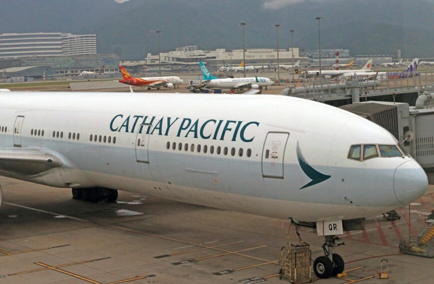 A Cathay Pacific airplane is parked at an airport gate. The aircraft is white with the airline's logo and name prominently displayed on the fuselage. In the background, several other airplanes from different airlines are visible on the tarmac, with airport buildings and mountains in the distance.