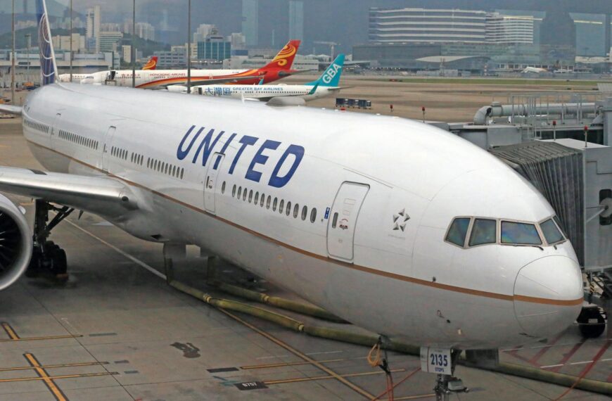 A large United Airlines airplane is parked at an airport gate, connected to a jet bridge. The aircraft is white with the airline's logo and name visible on the fuselage. In the background, other airplanes from different airlines are also parked, and airport buildings are visible under a cloudy sky.