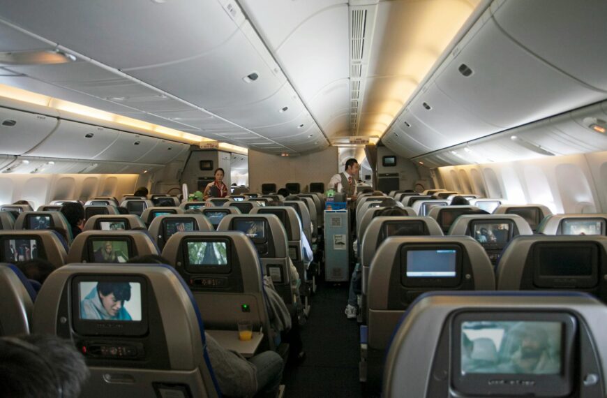 The image shows the interior of an airplane cabin, specifically the economy class section. There are multiple rows of seats, each equipped with personal entertainment screens. Passengers are seated, and two flight attendants are visible in the aisle, attending to the passengers. The cabin is well-lit, and some screens display various media content.