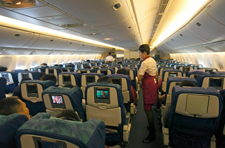 The image shows the interior of an airplane cabin with rows of seats. Each seat has a personal entertainment screen on the back. Passengers are seated, and a flight attendant in a red uniform is walking down the aisle. The cabin is well-lit, and the overhead compartments are closed.