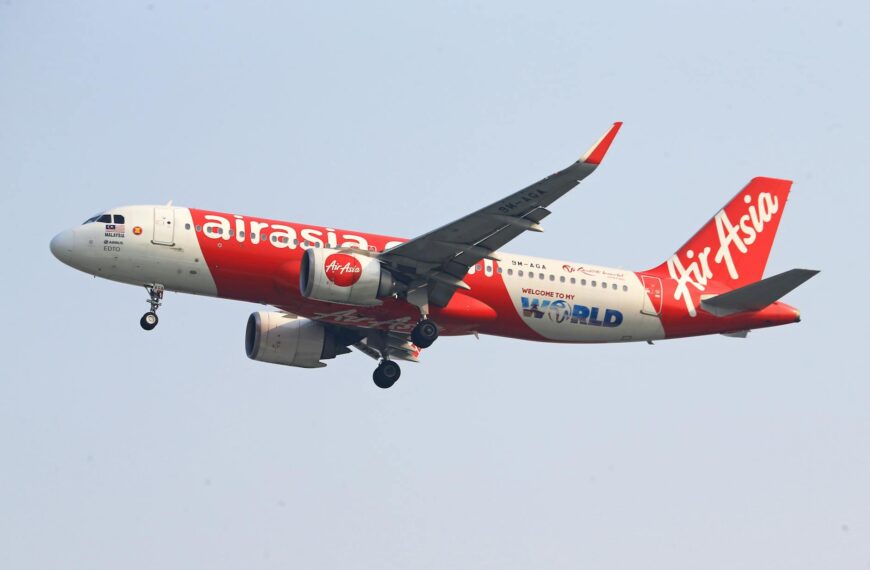 The image shows an AirAsia airplane in flight against a clear sky. The aircraft is predominantly red and white, with the AirAsia logo prominently displayed on the body and tail. The landing gear is extended, indicating that the plane is either taking off or preparing to land.