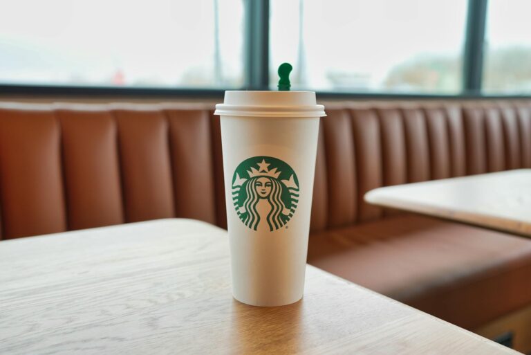A white Starbucks cup with a green logo is placed on a wooden table in a cafe setting. The background shows a brown cushioned bench and large windows.
