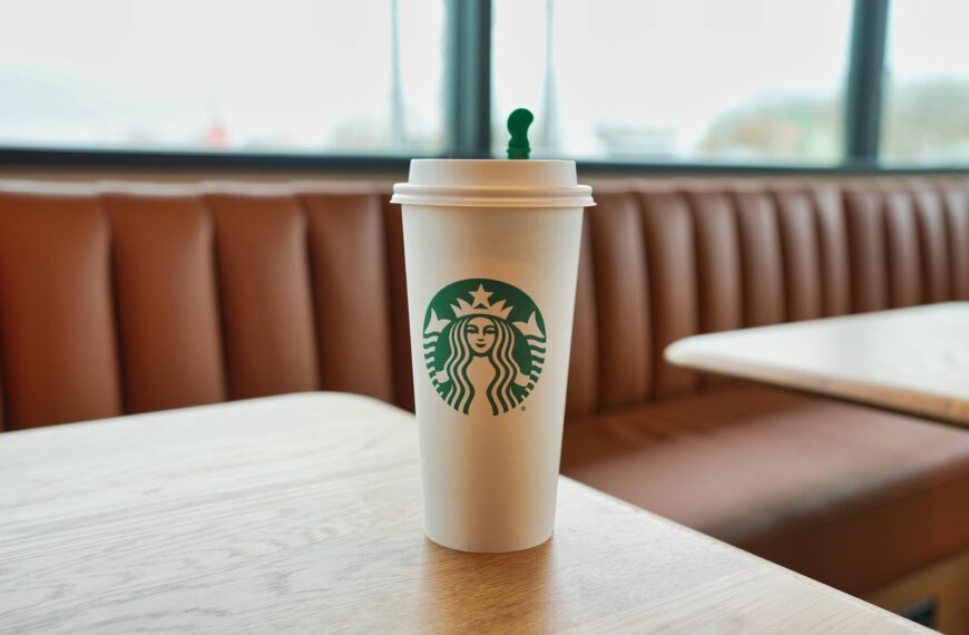 A white Starbucks cup with a green logo is placed on a wooden table in a cafe setting. The background shows a brown cushioned bench and large windows.