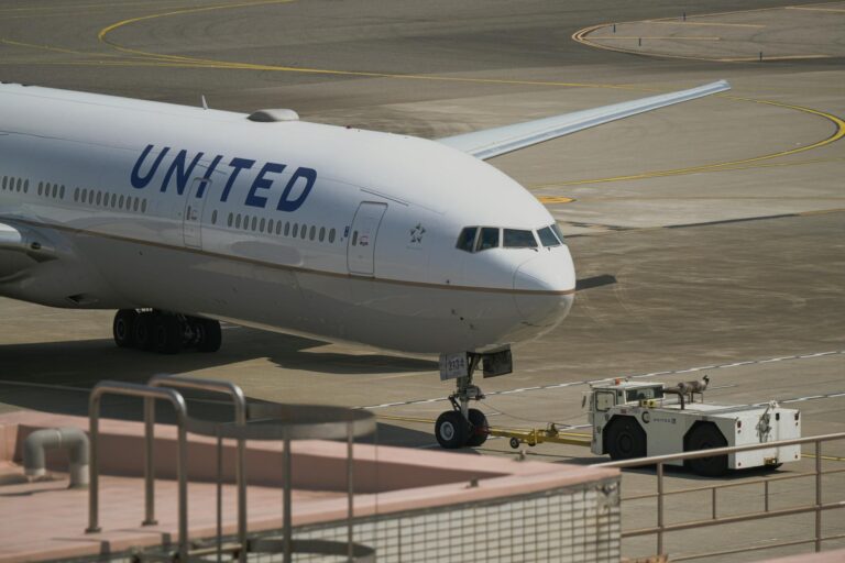 A United Airlines airplane is on the tarmac, being towed by a ground vehicle. The aircraft is positioned at an airport, with its nose facing the camera. The runway and taxiway markings are visible in the background.