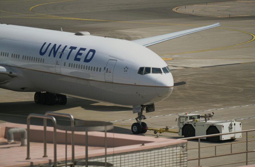 A United Airlines airplane is on the tarmac, being towed by a ground vehicle. The aircraft is positioned at an airport, with its nose facing the camera. The runway and taxiway markings are visible in the background.