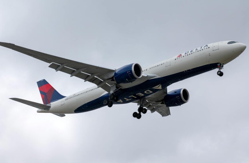 A large commercial airplane in flight against a cloudy sky. The aircraft has a white fuselage with blue and red accents, and the landing gear is extended. The airline's logo is visible on the body and tail of the plane.