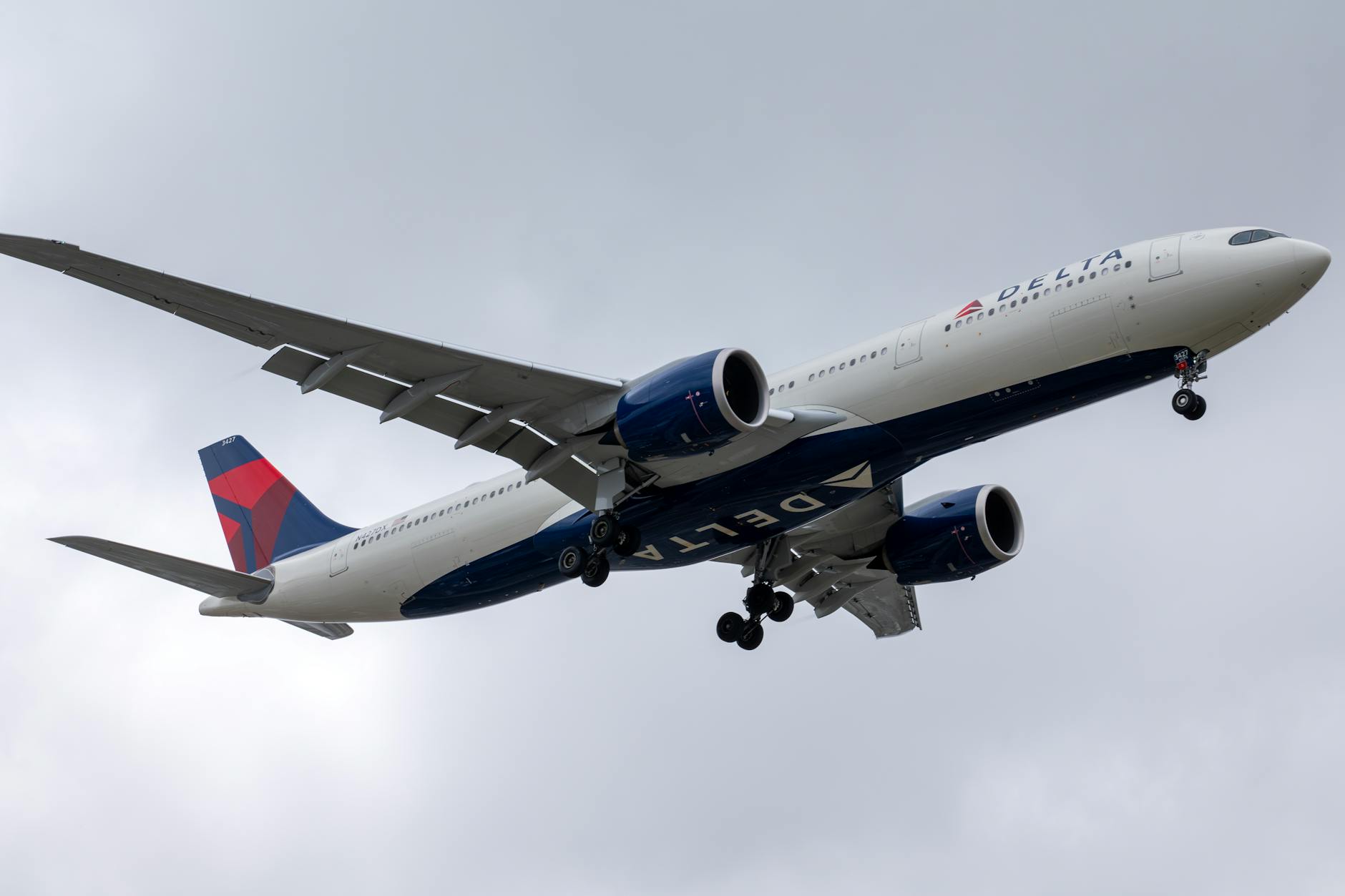 A large commercial airplane in flight against a cloudy sky. The aircraft has a white fuselage with blue and red accents, and the landing gear is extended. The airline's logo is visible on the body and tail of the plane.