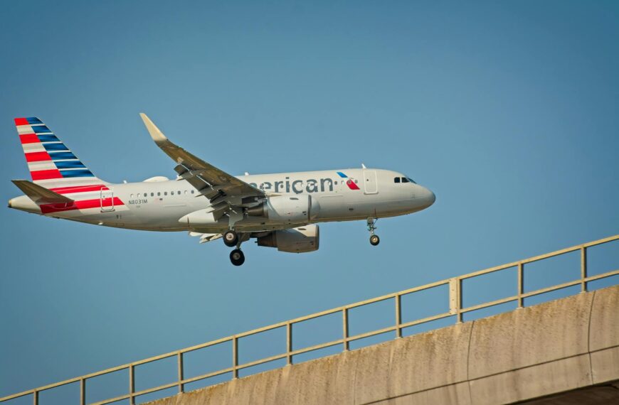 A commercial airplane with a silver body and a tail featuring red, white, and blue stripes is flying in the sky. The landing gear is deployed, and the plane is positioned above a concrete bridge. The sky is clear and blue.