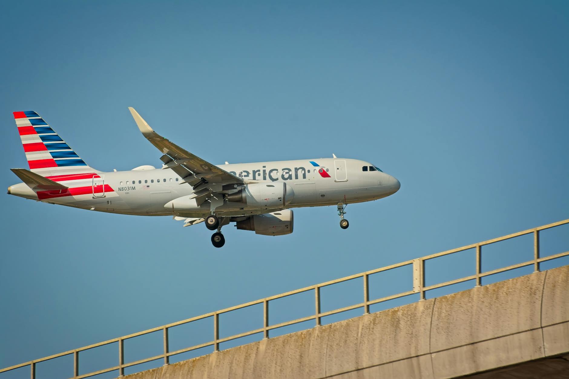 A commercial airplane with a silver body and a tail featuring red, white, and blue stripes is flying in the sky. The landing gear is deployed, and the plane is positioned above a concrete bridge. The sky is clear and blue.