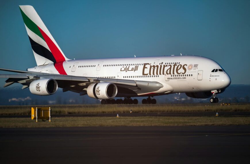 The image shows an Emirates Airbus A380 airplane on a runway. The aircraft is white with the Emirates logo and branding on the side. The tail fin features the airline's colors: green, red, and black. The plane is positioned on the tarmac with a clear blue sky in the background.