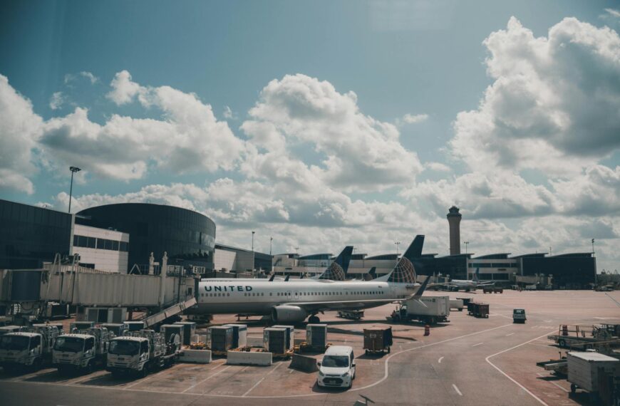 The image shows an airport scene with a United Airlines plane parked at a gate. The airport terminal is visible in the background, along with several jet bridges and service vehicles. The sky is partly cloudy, and an air traffic control tower is seen in the distance.