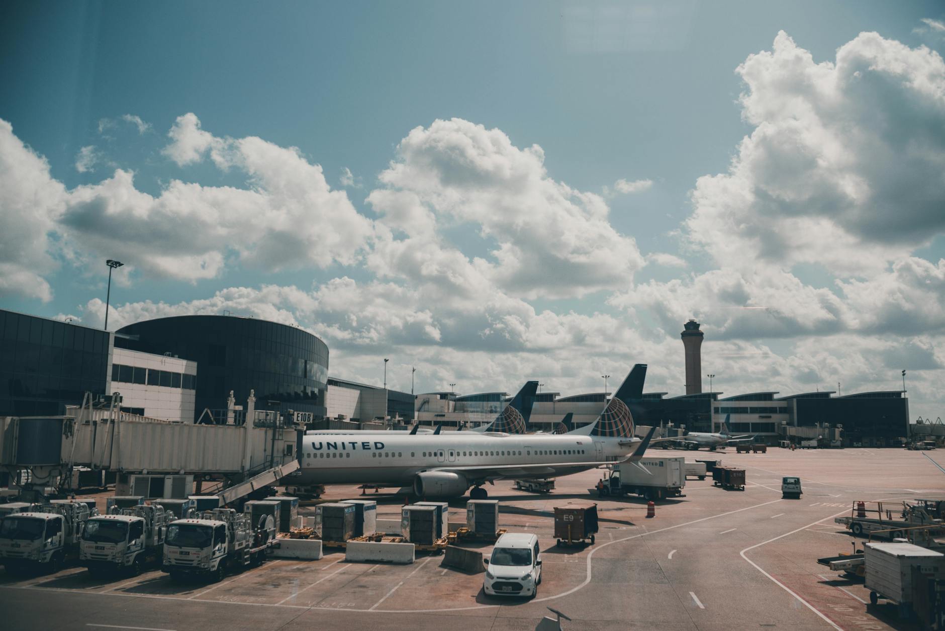 The image shows an airport scene with a United Airlines plane parked at a gate. The airport terminal is visible in the background, along with several jet bridges and service vehicles. The sky is partly cloudy, and an air traffic control tower is seen in the distance.
