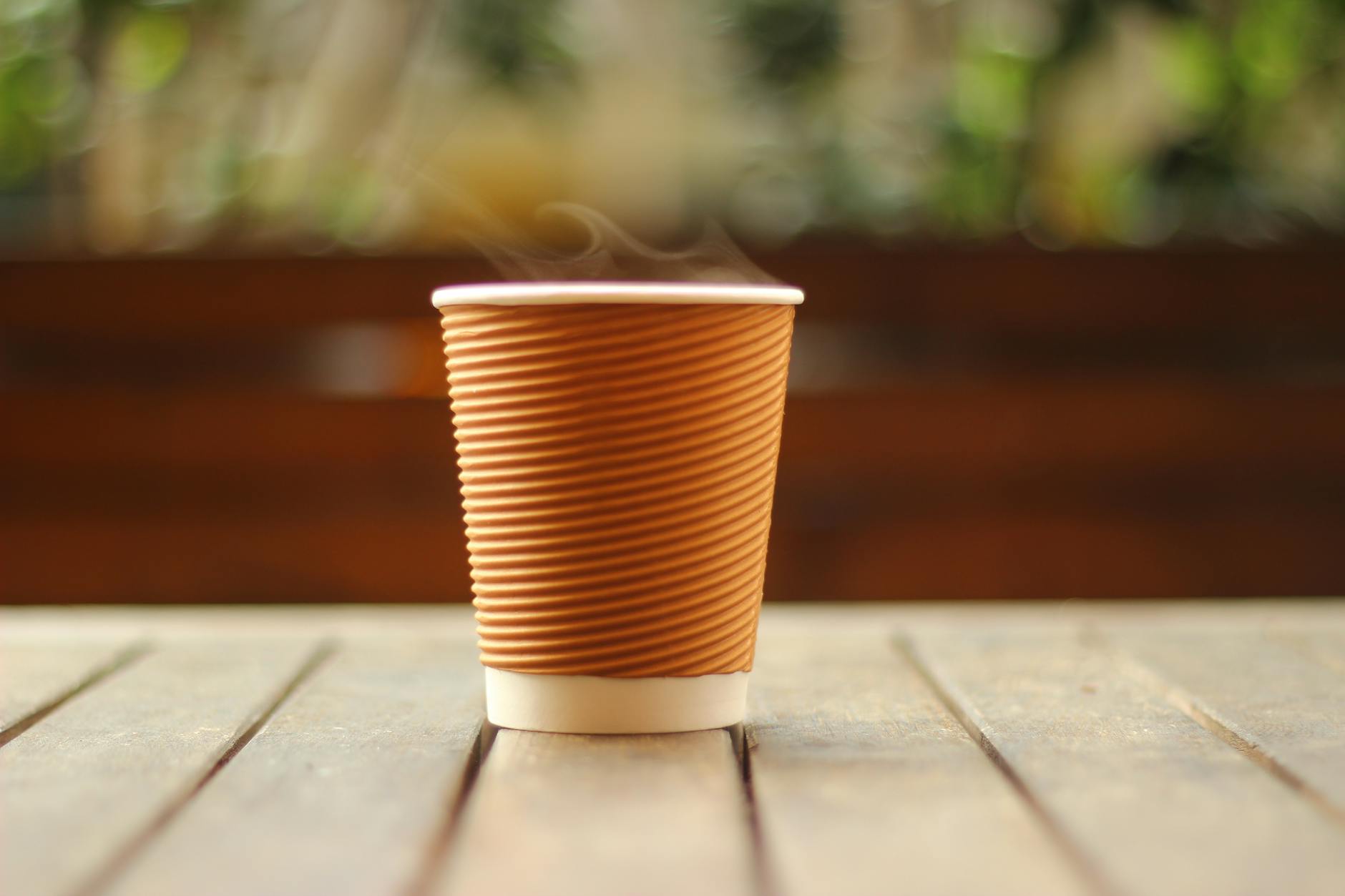 A close-up of a steaming hot coffee in a brown paper cup on a wooden table indoors.