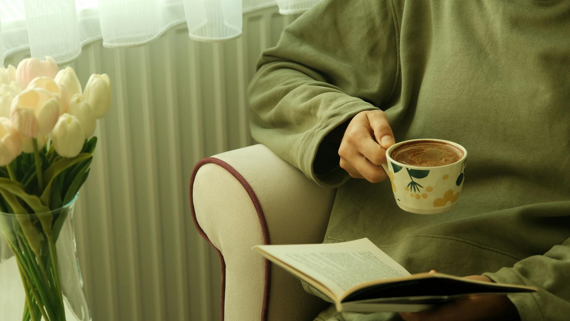 Person relaxing with coffee and book indoors, tulips beside them, cozy atmosphere.