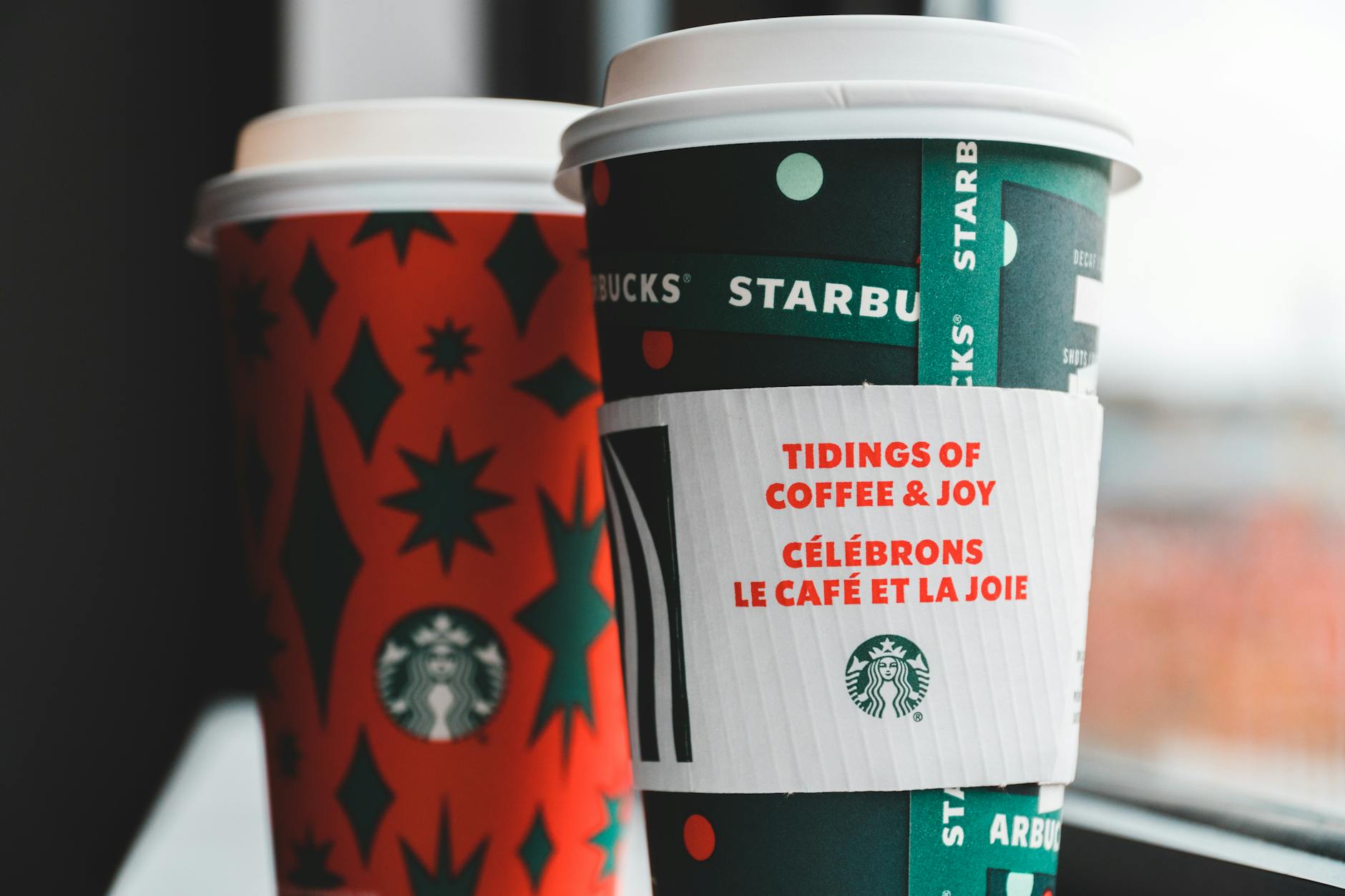 Close-up of festive Starbucks coffee cups with holiday-themed designs by a window.