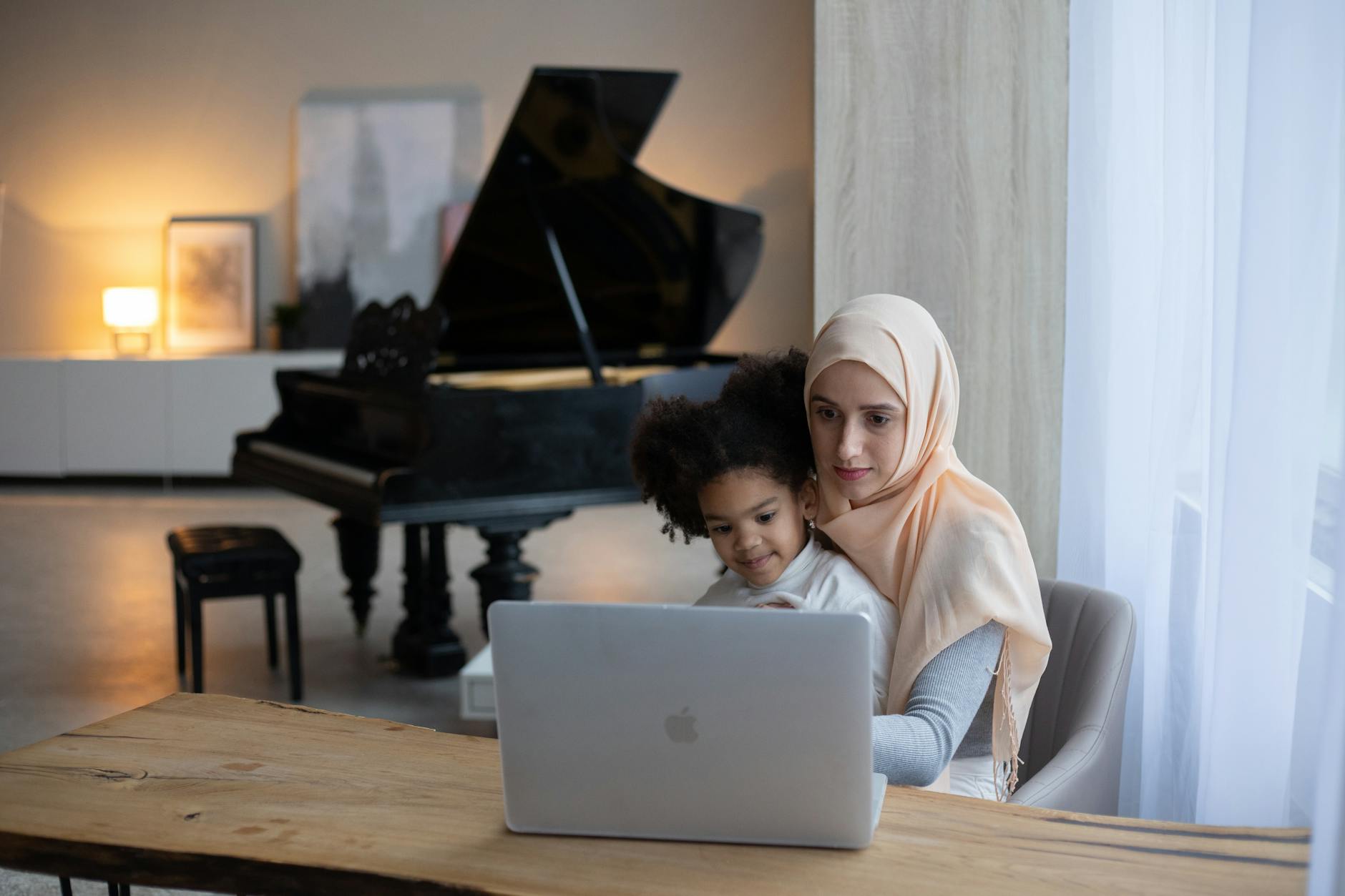 Friendly Muslim mother sitting with little African American daughter on knees and watching cartoon on portable laptop in cozy apartment